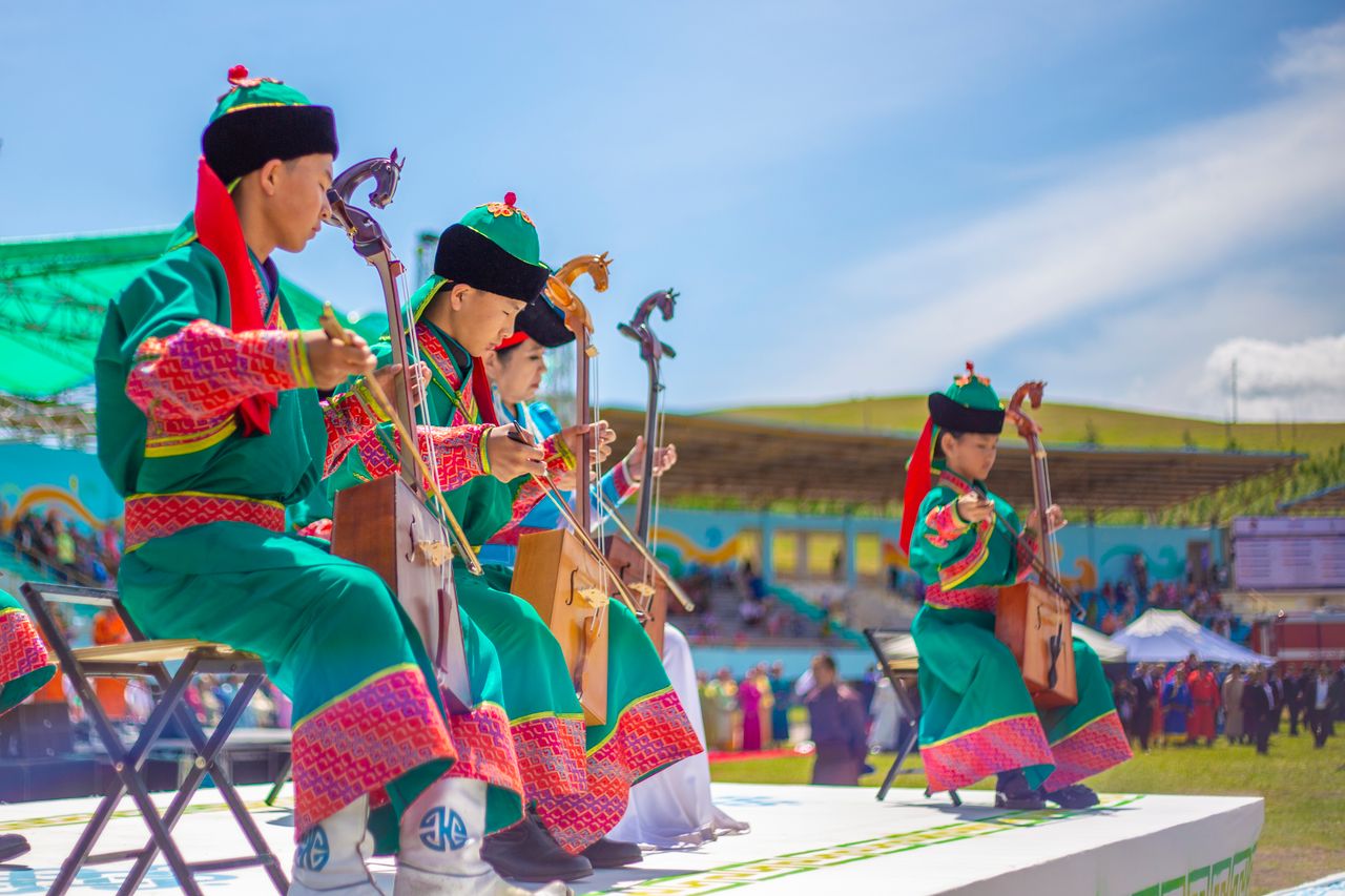 gallery-1 Four musicians in colorful Mongolian dress perform with traditional instruments outdoors.