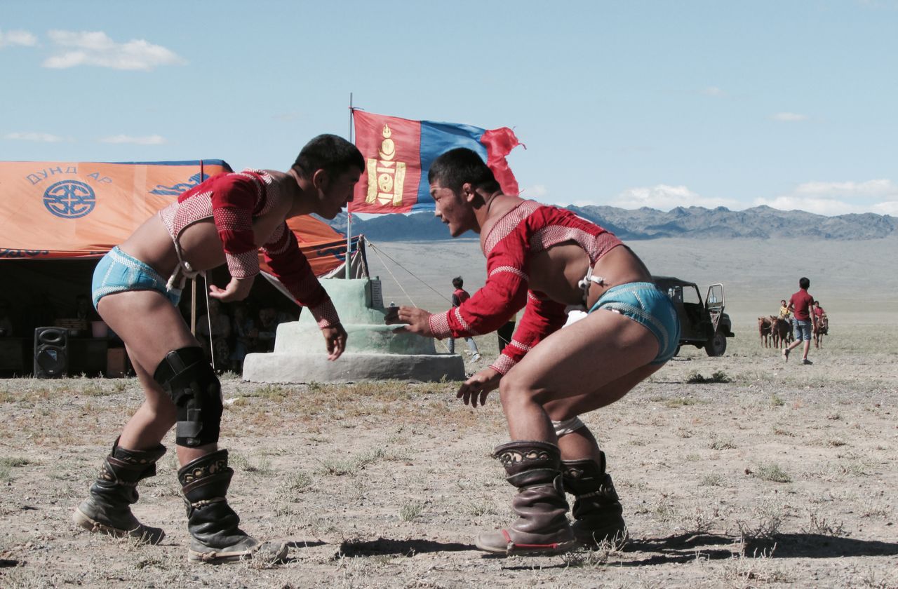 gallery-3 Two men compete in traditional Mongolian wrestling at a festival outdoors with cultural elements visible.